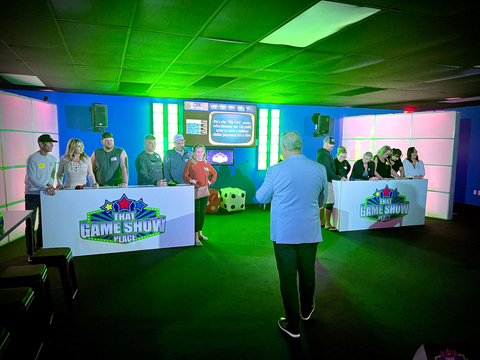 Two teams at podiums facing the host in the game show studio, scoreboard lit behind them