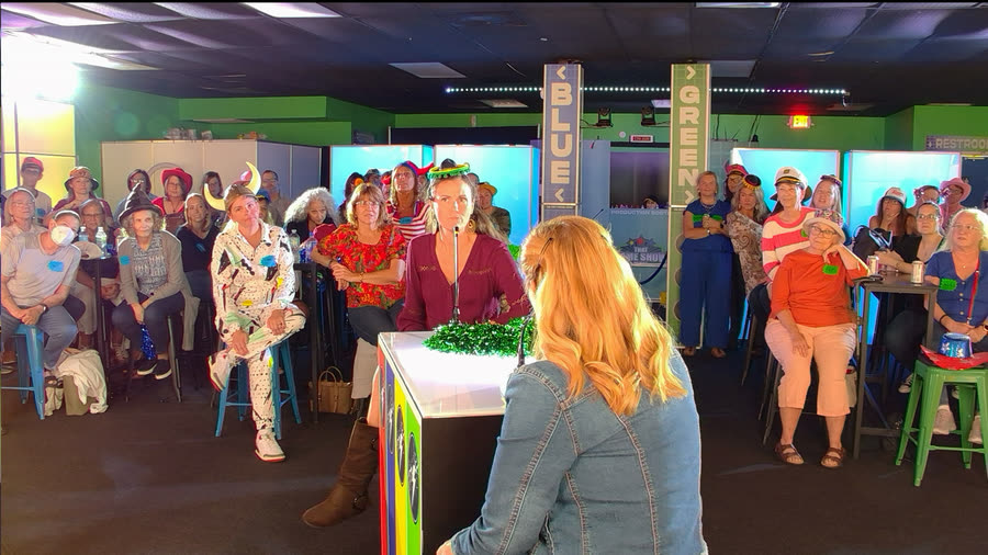 The host at a lit podium addressing a seated audience in party hats