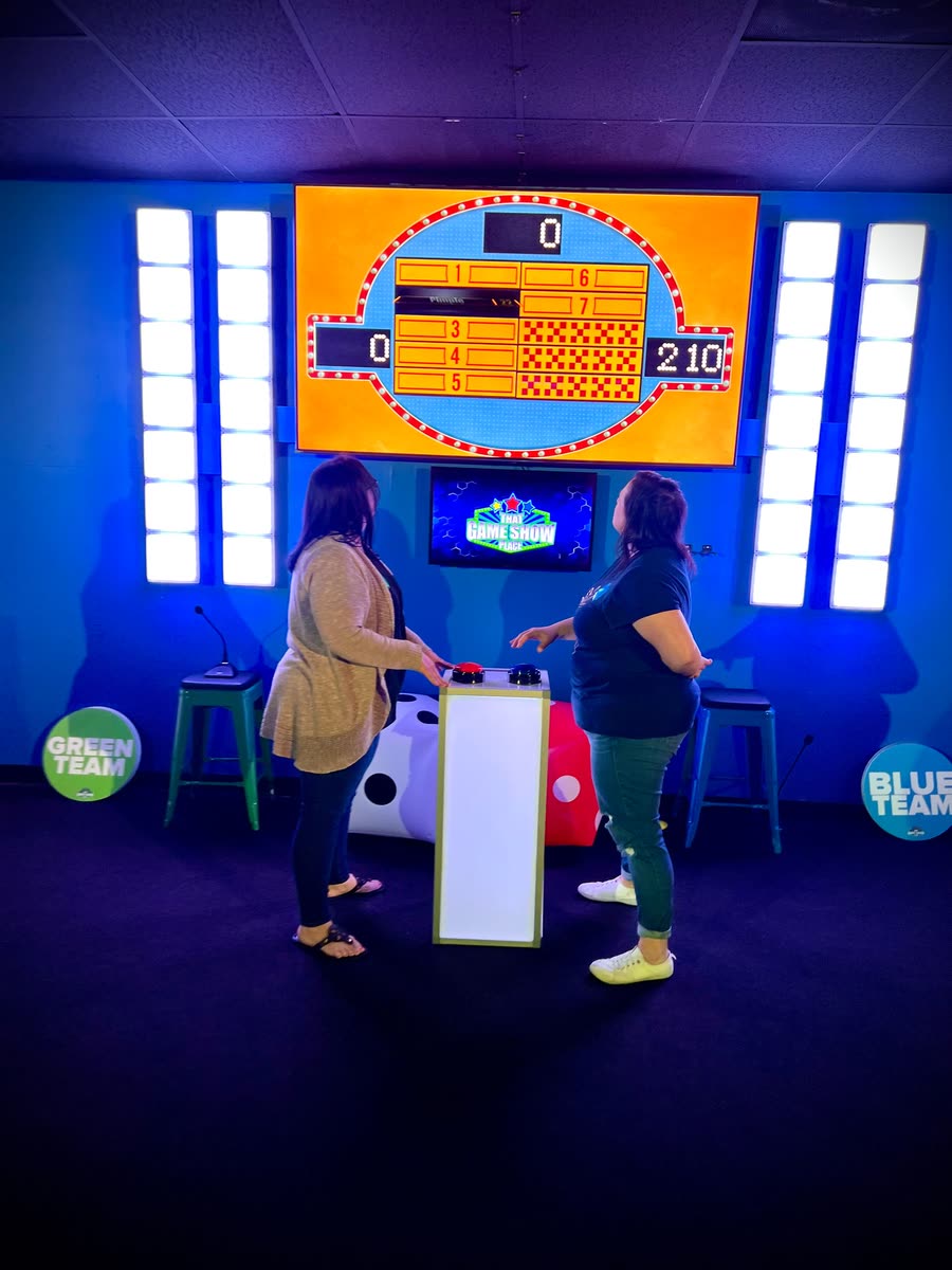 Two players facing off at a buzzer podium below a lit scoreboard, Green Team and Blue Team signs on the wall