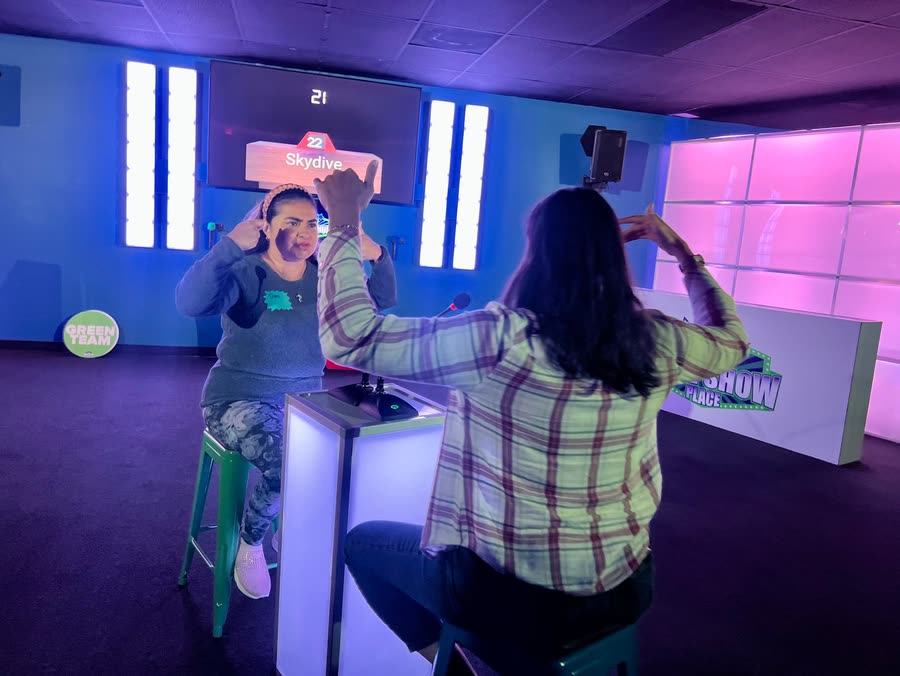 A player at a lit podium acting out a clue while a teammate guesses
